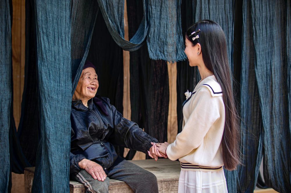 An elder and a young woman clasping hands among indigo-dyed cloths